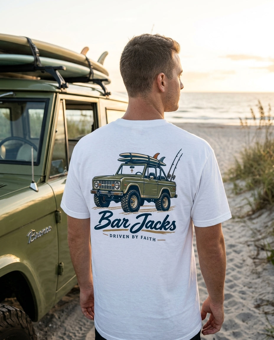 Man wearing the Bar Jacks Driven By Faith white cotton tee beside a vintage 4x4 at a beach dune crossover, back print visible