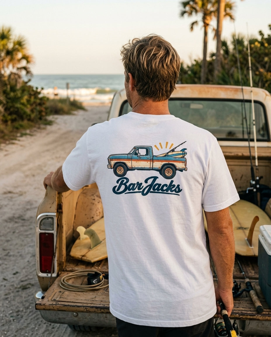 Man wearing the Bar Jacks Vintage Beach Truck white cotton tee at a pickup tailgate on a beach road, back print visible